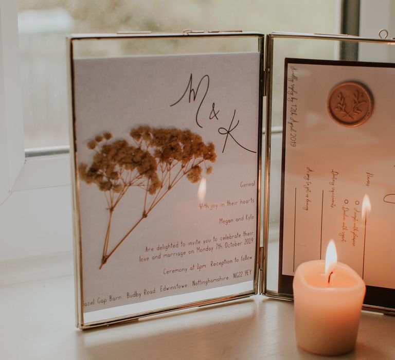 Pressed flower and wax seal invites in golden frame next to candle for barn wedding ceremony