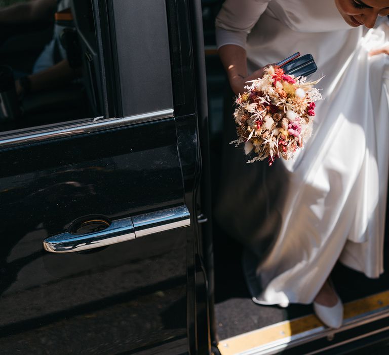 Bride leaves car wearing Etsy gown from PionDress as she holds vibrant dried flower bouquet in one hand and lifts skirt in the other