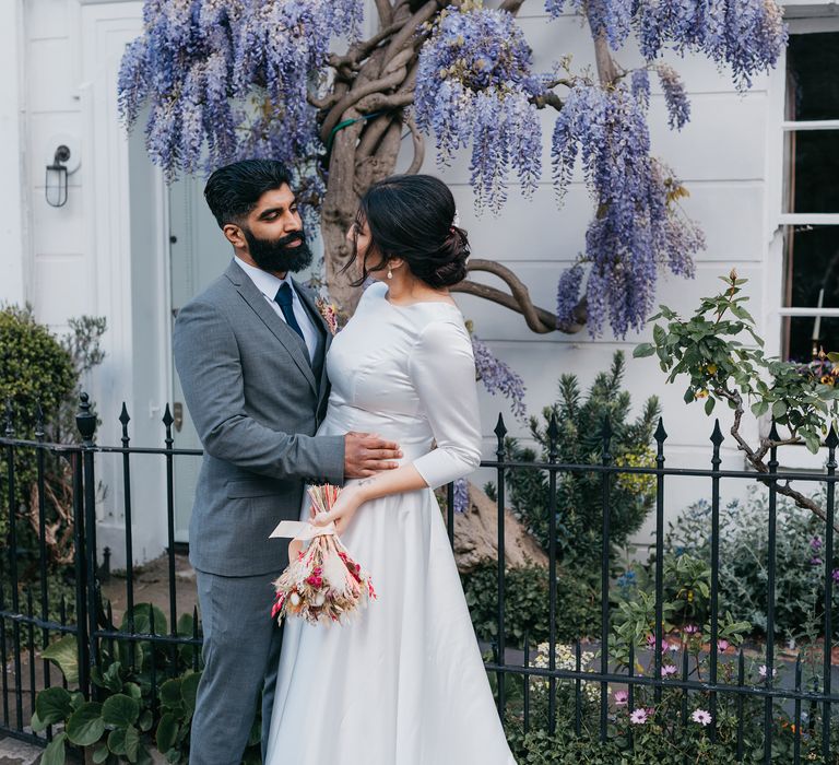 Bride & groom embrace outside house with purple wisteria tree outside and black iron fence