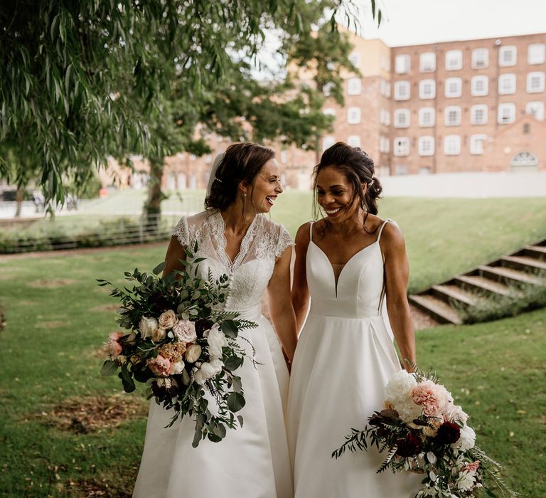 Bride in white cami wedding dress stands with bride in white lace top capped sleeve wedding dress as they both laugh holding white, pink and green bridal bouquets outside The West Mill Derby