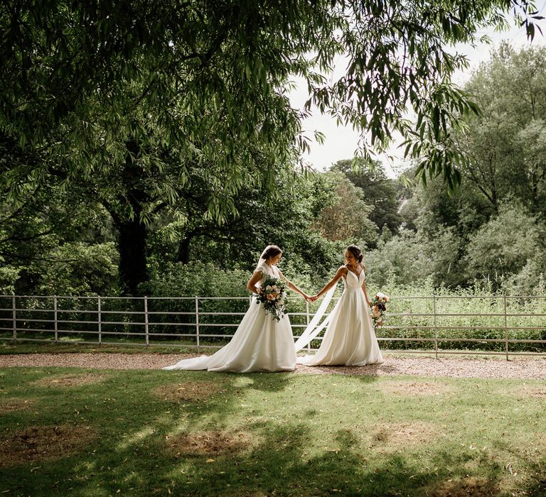 Bride in white cami wedding dress and shoulder veils walks through the grounds of The West Mill Derby holding hands with bride in lace top capped sleeve wedding dress and veil 