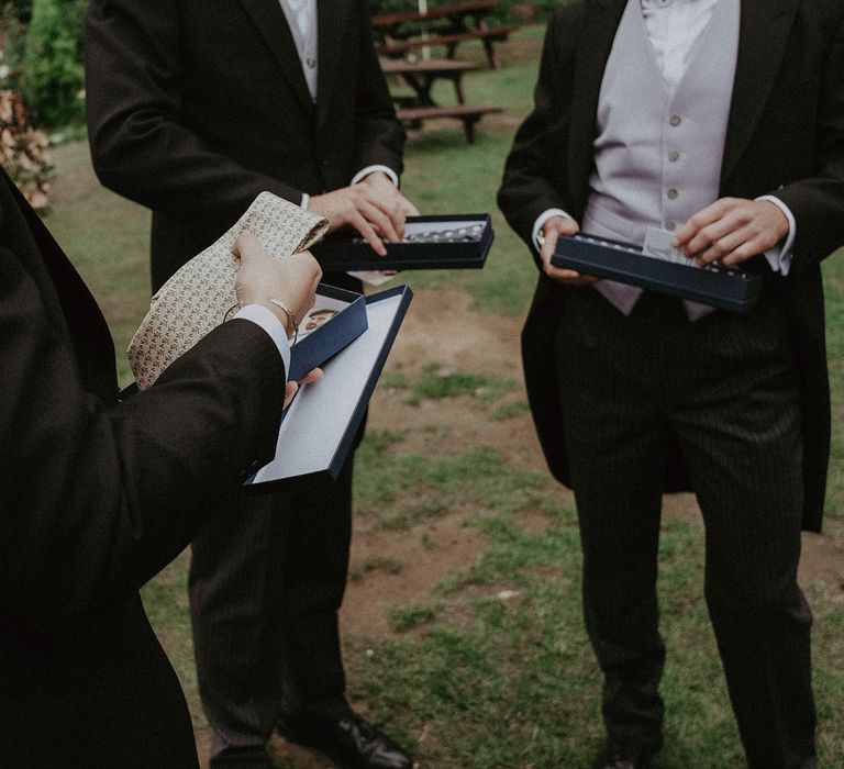 Groom and groomsmen stand in garden looking at their bespoke Marmaduke ties with olive leaf motif before Surrey summer wedding