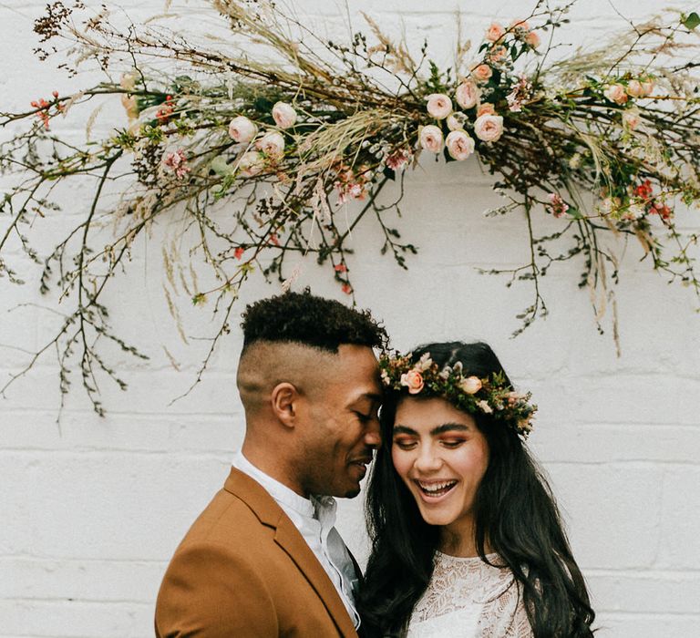 Groom in a bronze coloured suit embracing his bride in a lace wedding dress and flower crown under a twig and pink flower installation