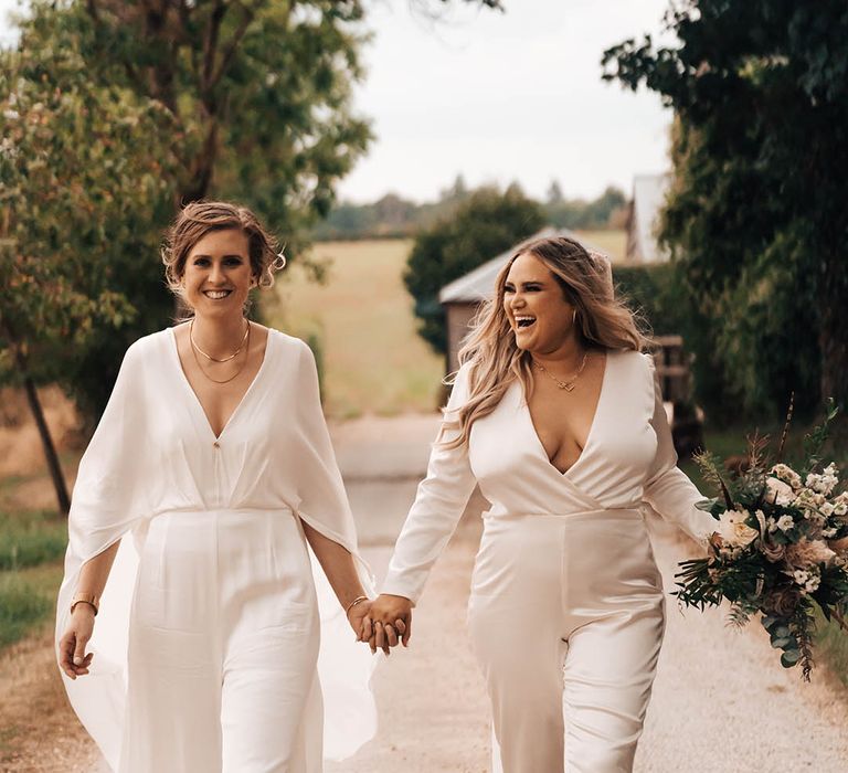 Brides in jumpsuits walk together down country lane hand in hand on their wedding day