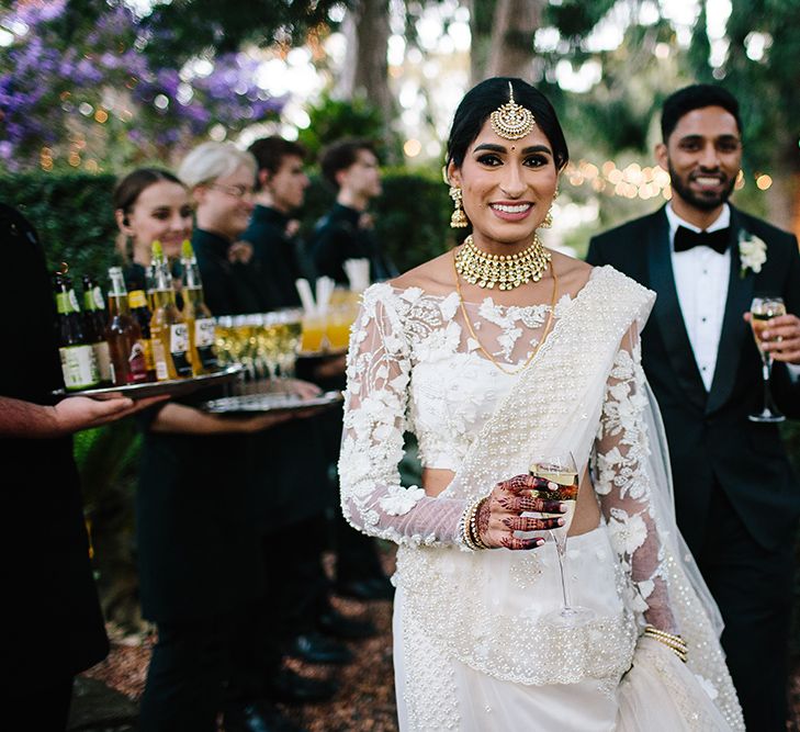 An Indian bride wears a white sari and gold jewellery. Her hair is tied back and she holds a glass of champagne.