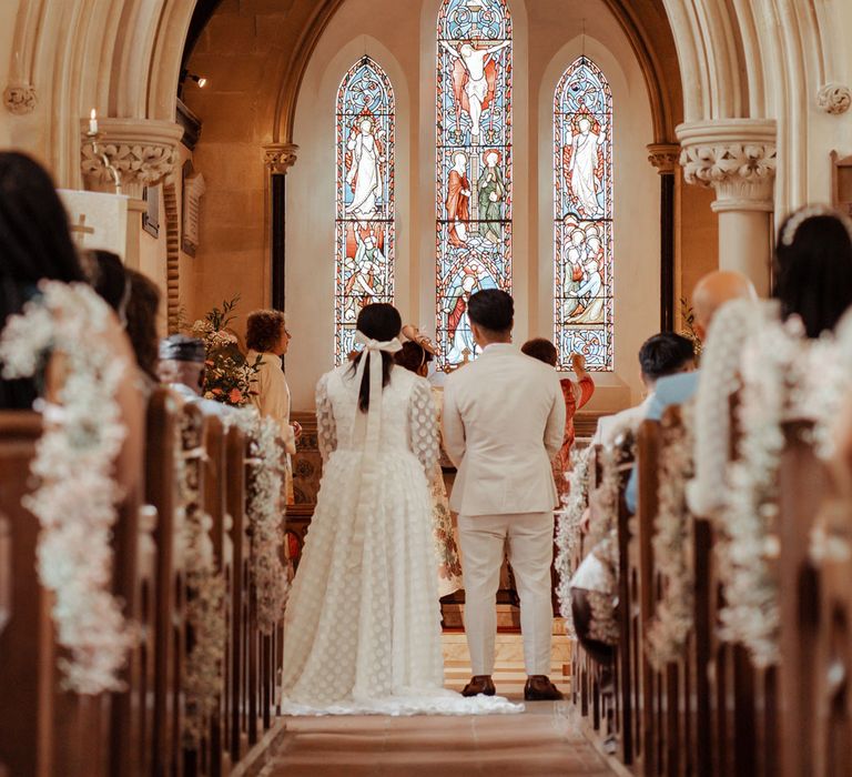 Bride in long mesh sleeve spotted Vagabond wedding dress and bridal hair bow stands with groom in linen suit at the altar in church during Wasing Park wedding