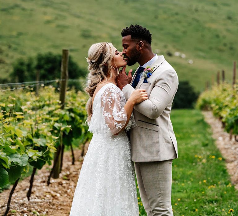 Groom in a beige suit with blue check kissing his bride in lace wedding dress at Little Wold Vineyard 