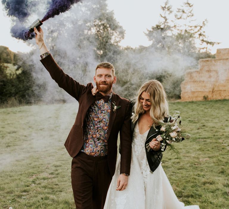 Bride in a tulle wedding dress and leather jacket holding onto her grooms arm in a floral shirt and tweed jacket as he holds a smoke bomb in the air 