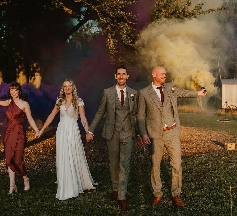 Bride in lace top capped sleeve wedding dress holding hands with groom in grey herringbone suit and bridesmaid in one shoulder burgundy satin bridesmaid dress as they walk next to groom holding smoke bomb during late summer wedding at Wellington Wood in Norfolk