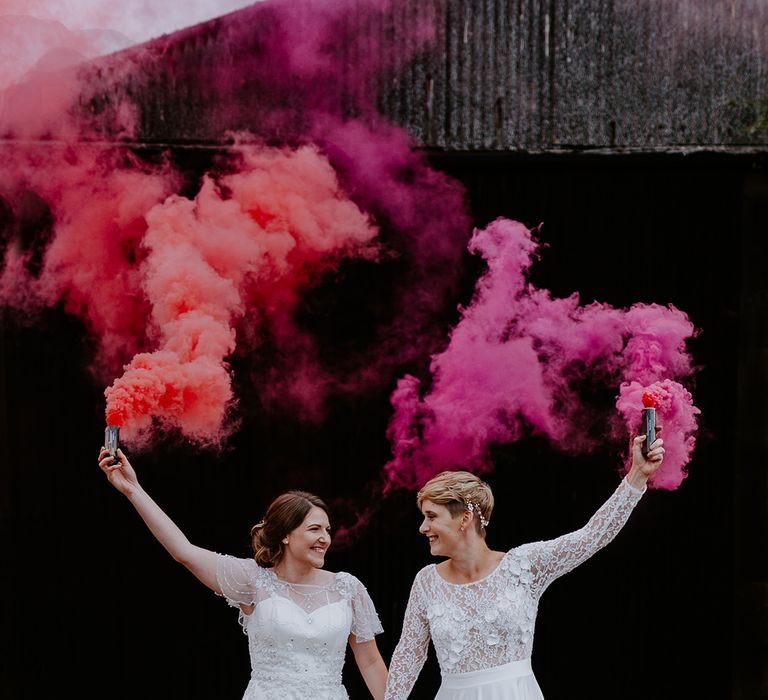 Portrait of two brides holding pink and red smoke bombs in the air at their Woolas Barn wedding 