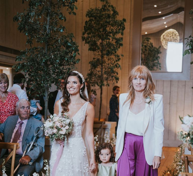 Bride in applique Berta wedding dress, headband and veil with white, blush pink and green bridal bouquet walks down the aisle with flower girl in sage green satin dress and women in purple satin trousers, white top and white blazer during wedding ceremony at Primrose Hill Farm