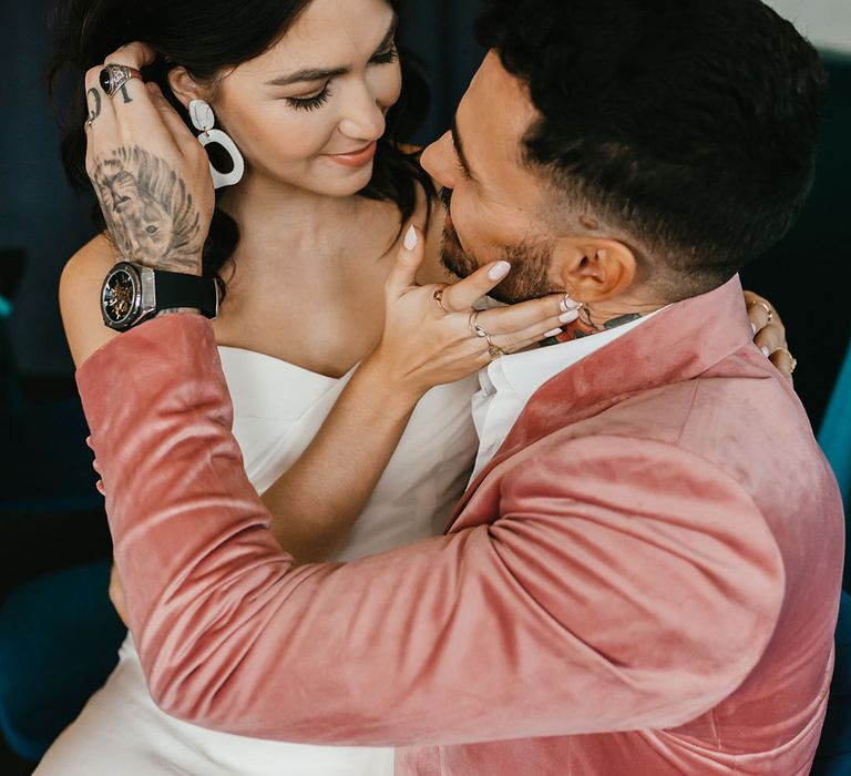 Groom in a dusky pink velvet jacket brushing his brides hair behind her ears to show off her ceramic earrings 