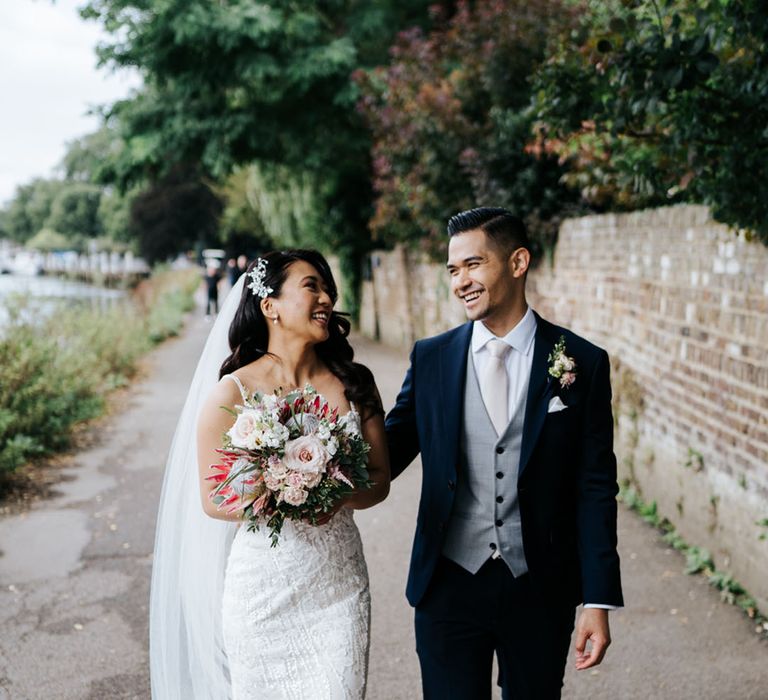 Bride in sparkly dress & Groom in simple suit with a waistcoat laughing as they walk under stunning greenery settings