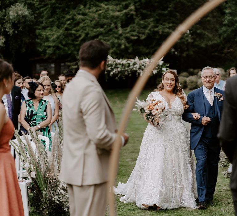 Bride in homemade wedding dress holding rose bridal bouquet walks up the aisle holding arm of man in blue three piece suit during DIY garden wedding ceremony in Bedfordshire