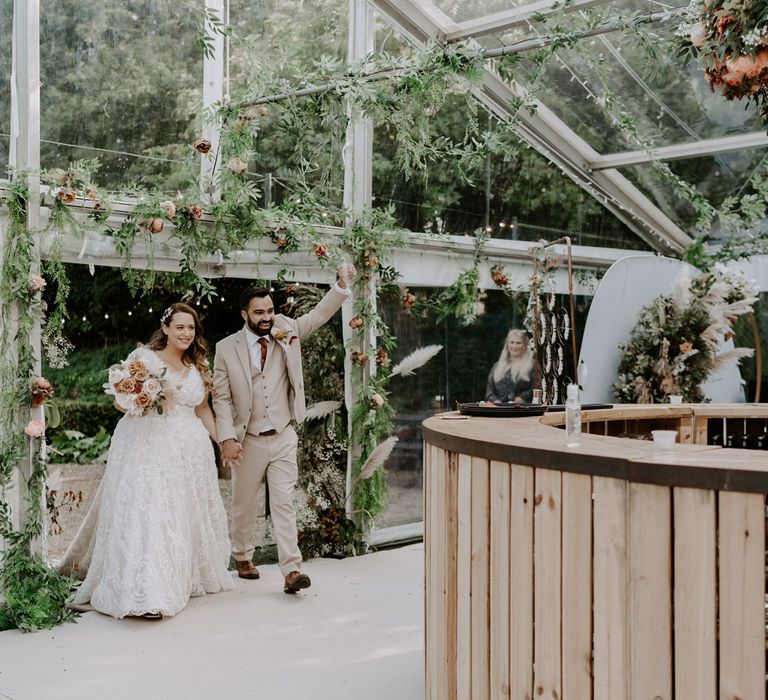 Bride in homemade lace wedding dress holding mixed rose bridal bouquet walks into marquee decorated with garlands holding hands with groom in three piece beige suit for garden wedding reception