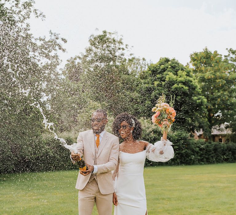 Stylish bride in a strapless wedding dress with long sheer sleeves and groom in a stone coloured suit popping champagne on the lawn 