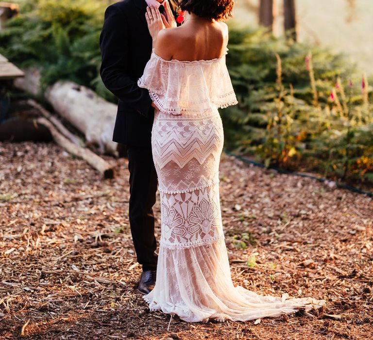 Bride in off the shoulder Grace Loves Lace wedding dress with train and colourful flower crown stands with hands on grooms chest in woodland during golden hour