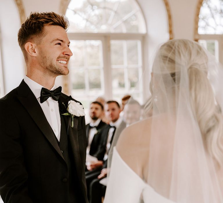 Excited groom in a tuxedo with white buttonhole flower holding hands with his bride at the altar 