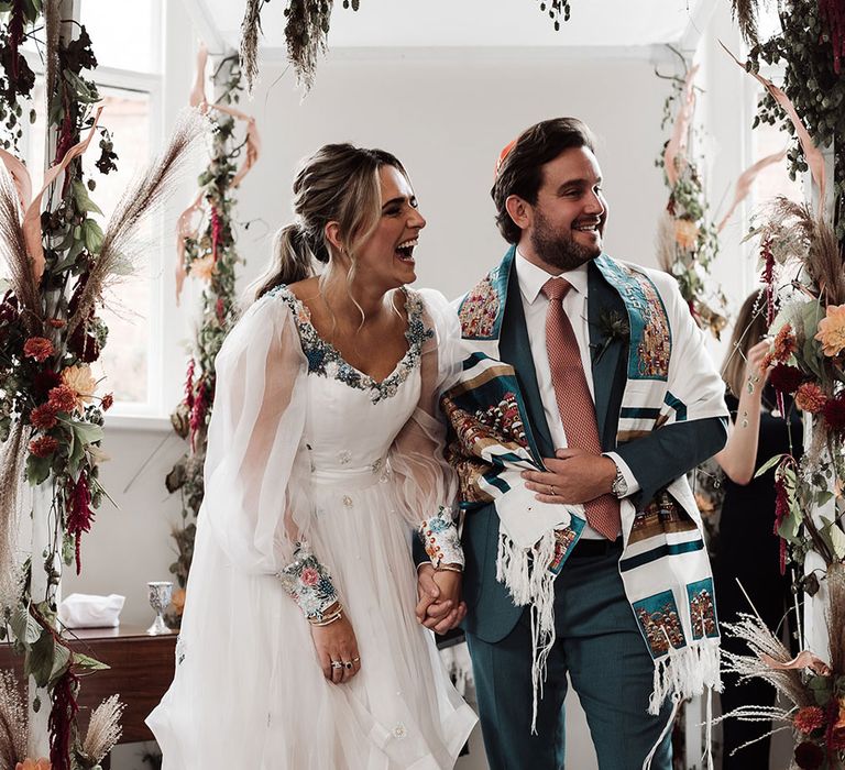 Bride in mesh sleeve embroidered Victoria Sanders wedding dress with train stands holding hands with groom in green suit, pink tie and tallit during interfaith wedding ceremony at Iscoyd Park