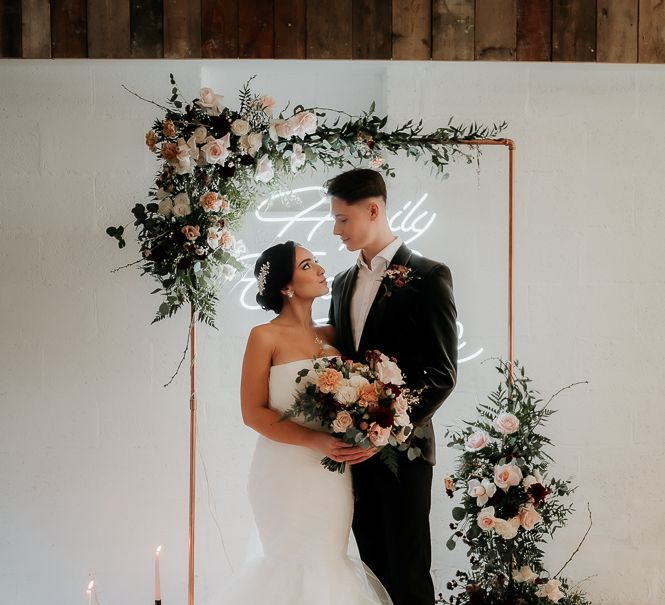 Bride looks up lovingly at groom in front of metal archway complete with floral decor and candle lights below 