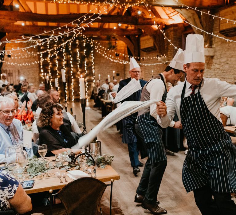 Groom and guests in striped aprons and chefs hats dance with tea towels down the aisle of barn wedding breakfast 