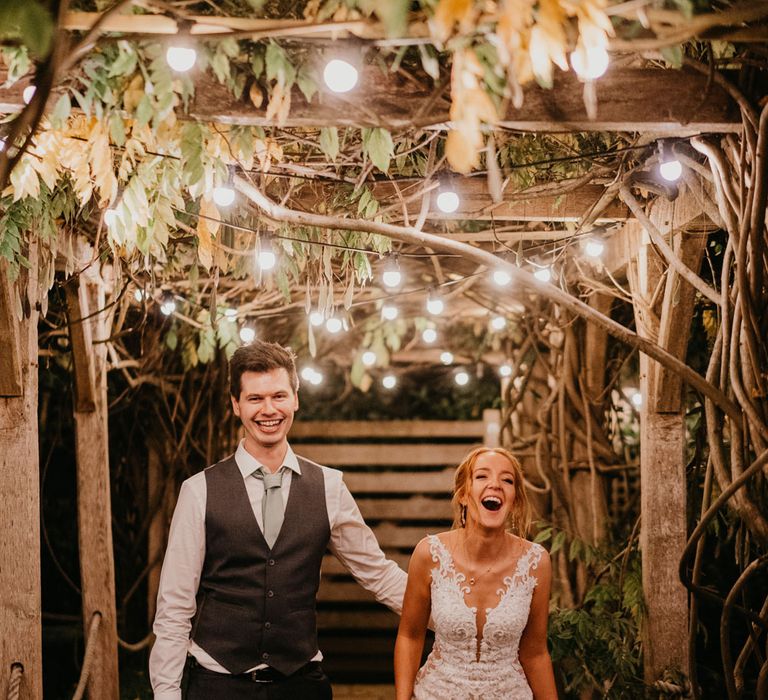 Bride in lace and tulle wedding dress stands with groom in white shirt and waistcoat on garden path lit by Edison bulbs after rustic barn wedding ceremony at The Tythe Barn in Launton