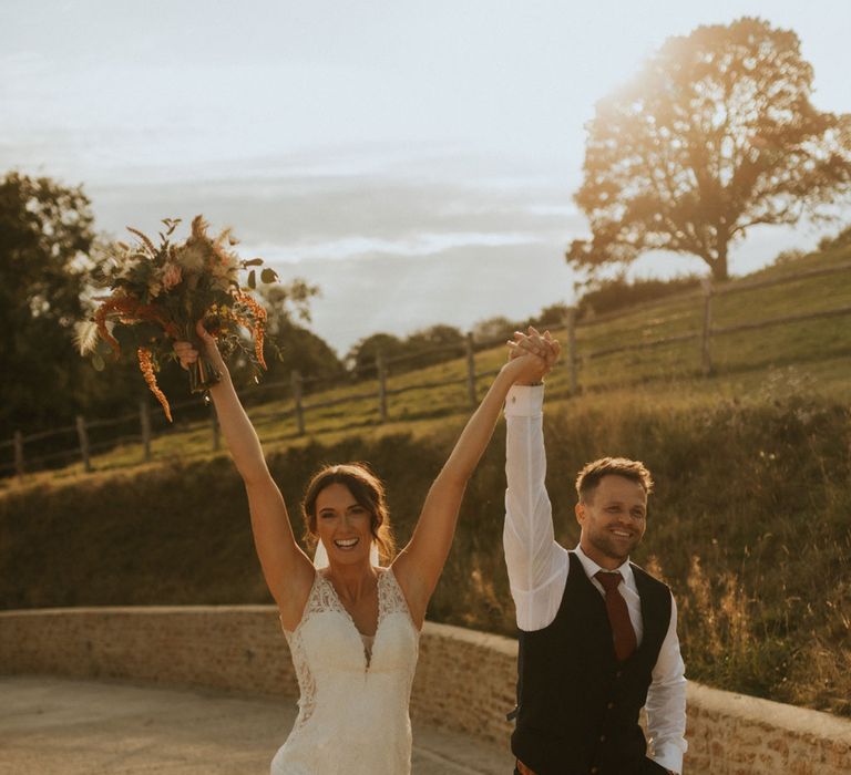 Bride in v neck lace wedding dress with train walks holding up hands with groom in white shirt and waistcoat at farm wedding in Dorset