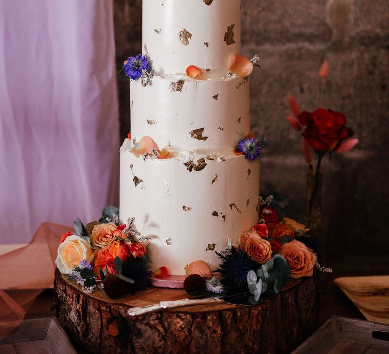 White iced three tier wedding cake with gold leaf detailing and floral decor for festival inspired wedding