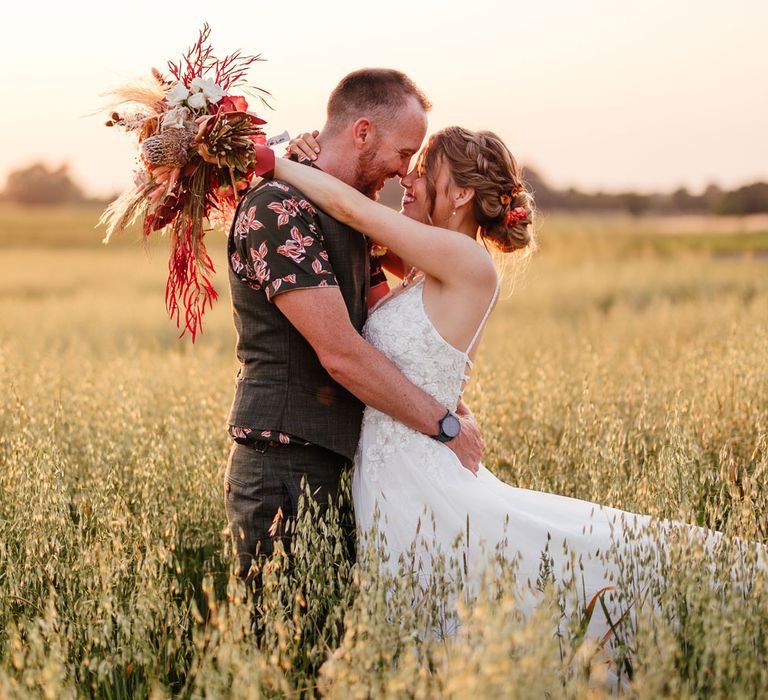 Bride in lace wedding dress with corset back holding dried floral bouquet hugs groom in short sleeve patterned shirt and waistcoat in field during golden hour