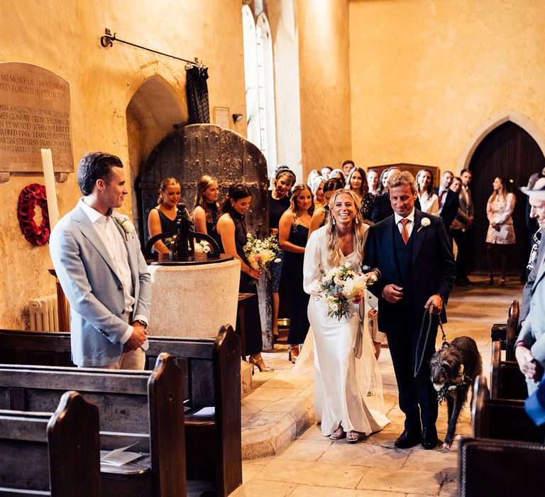 Bride in white satin dress holding mixed bridal bouquet walks down the aisle with man in three piece suit and dog in floral collar on a lead for church wedding ceremony in Hampshire 