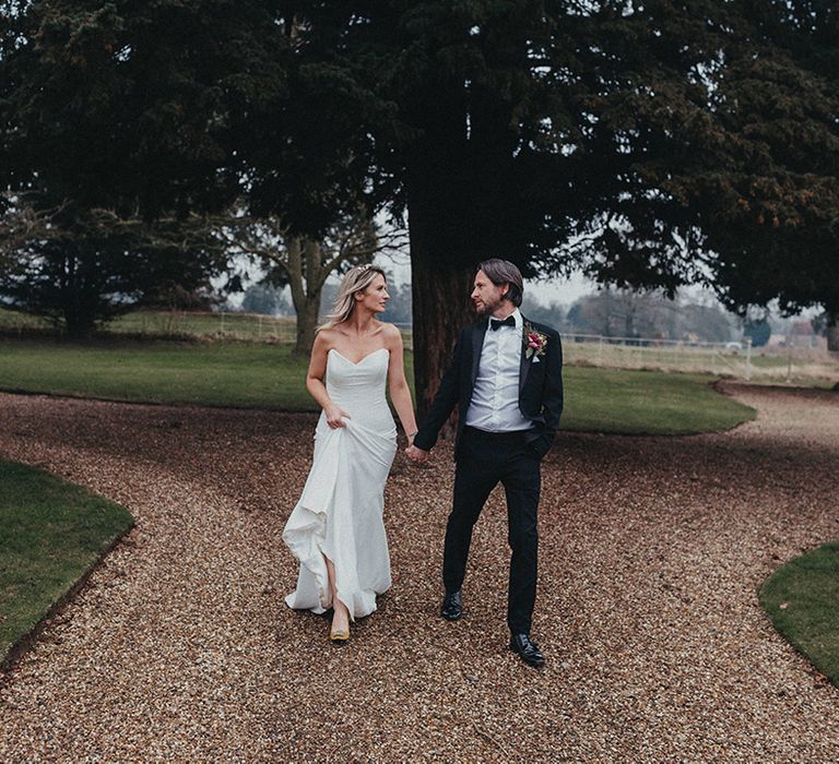 Bride holds the train of her Suzanne Neville gown as she walks with her groom who wears black tie 