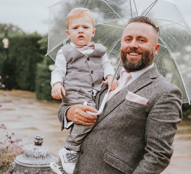 Groomsman with mini groomsman under clear umbrella on rainy day at country house wedding venue