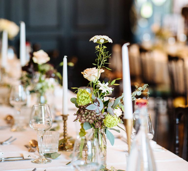 Tablescape with white candles in antique gold candleholders with milk bottle vases holding assortment of greenery and roses 