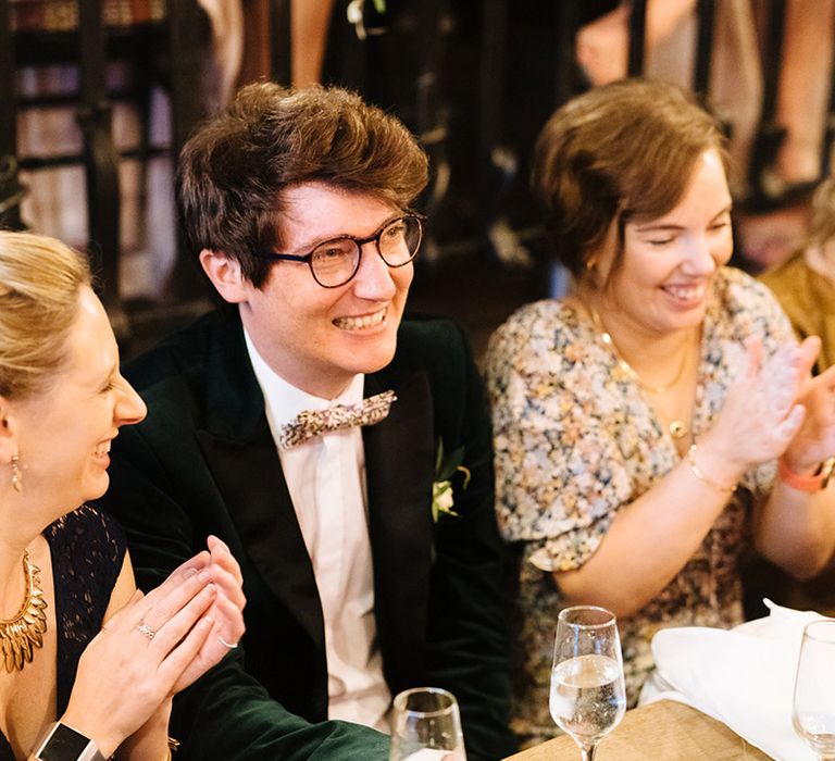 Groom in green velvet tuxedo sits with wedding guests at wedding reception