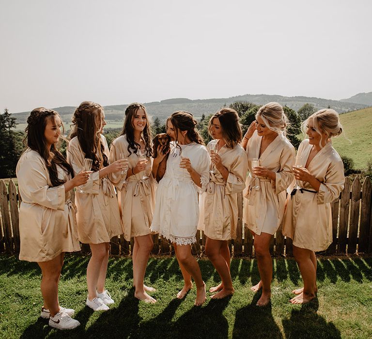 Bride holding dachshund dog in lace satin robe with bridesmaids in gold robes getting ready for the wedding