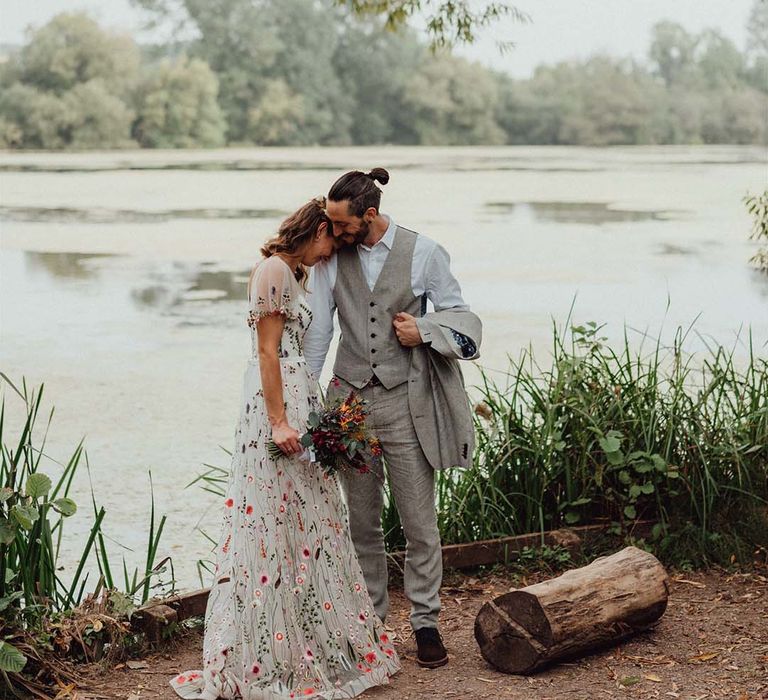 Bride in intricate floral embroidered wedding dress with groom in grey outfit stand close together at lake wedding venue 