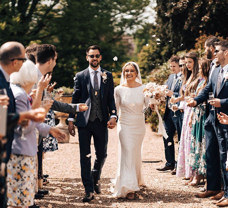 Groom in black sunglasses and dark blue suit with bride walks through their wedding guests throwing confetti