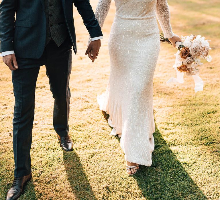 Bride in long sleeve beaded wedding dress walking and holding hands with groom in blue suit with paisley tie