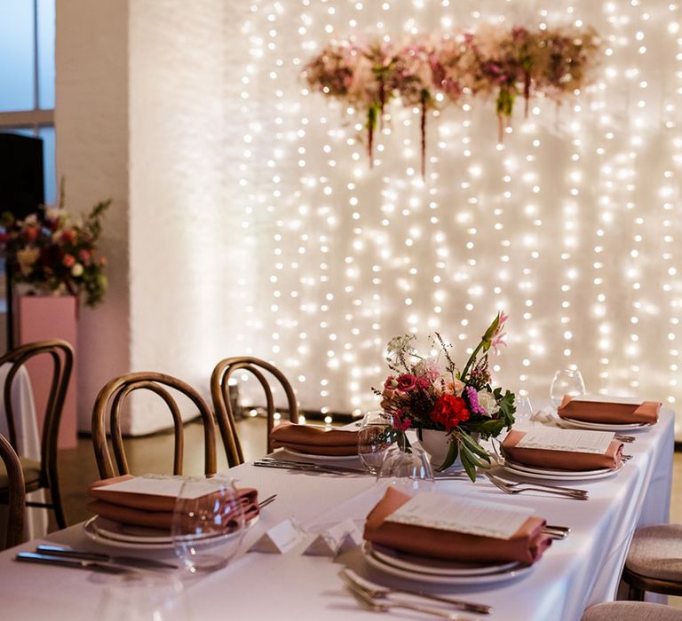 Wedding table settings with orange napkins and bright pink flowers with fairy lights and dried flower cloud for decor 