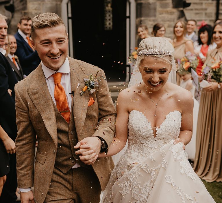 Bride in strapless sweetheart lace wedding gown with pearl tiara has confetti exit with groom in brown and orange suit and tie 