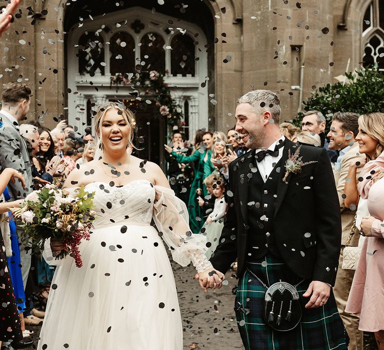 Groom smiles at the bride as she smiles at the camera as they walk through black confetti 