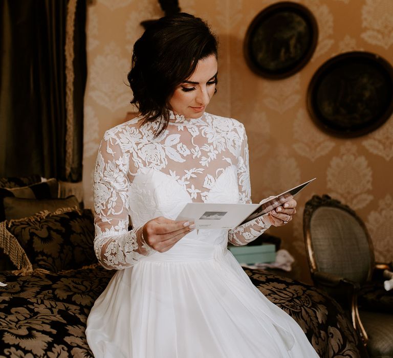 Bride in sweetheart illusion lace wedding dress with long sleeves reads a card on the morning of the wedding