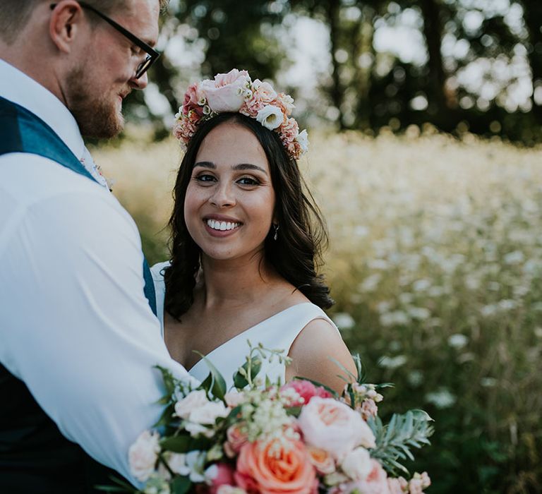 Bride with curled dark hair wears a pink flower crown holding a pink bouquet with groom in blue waistcoat holding her 