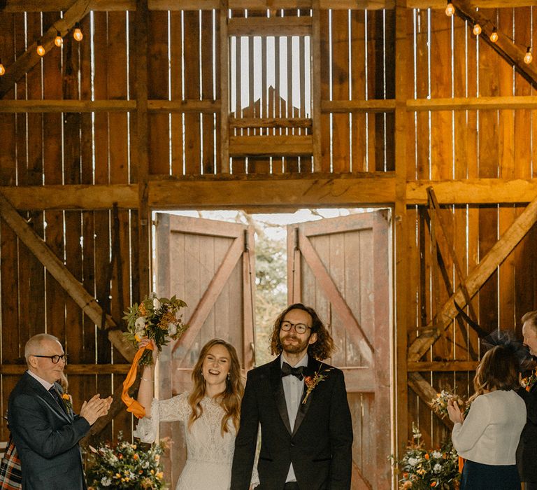 Bride and groom exit their wedding ceremony as a married couple with bride lifting autumnal bouquet with orange ribbon