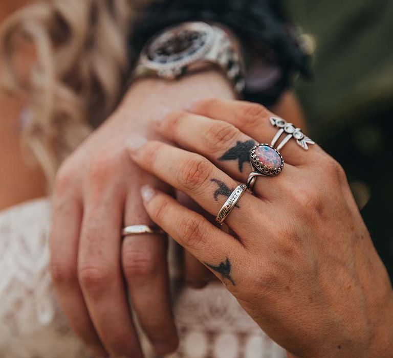 Bride wearing multiple silver rings on tattooed hand including engraved wedding band holding onto groom's hand with silver wedding band and watch