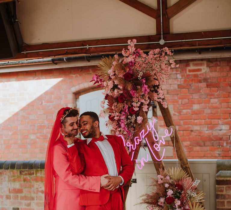 LGBTQI+ couple standing at the wooden frame altar decorated with pink flowers and dried grasses in coloured groom suits