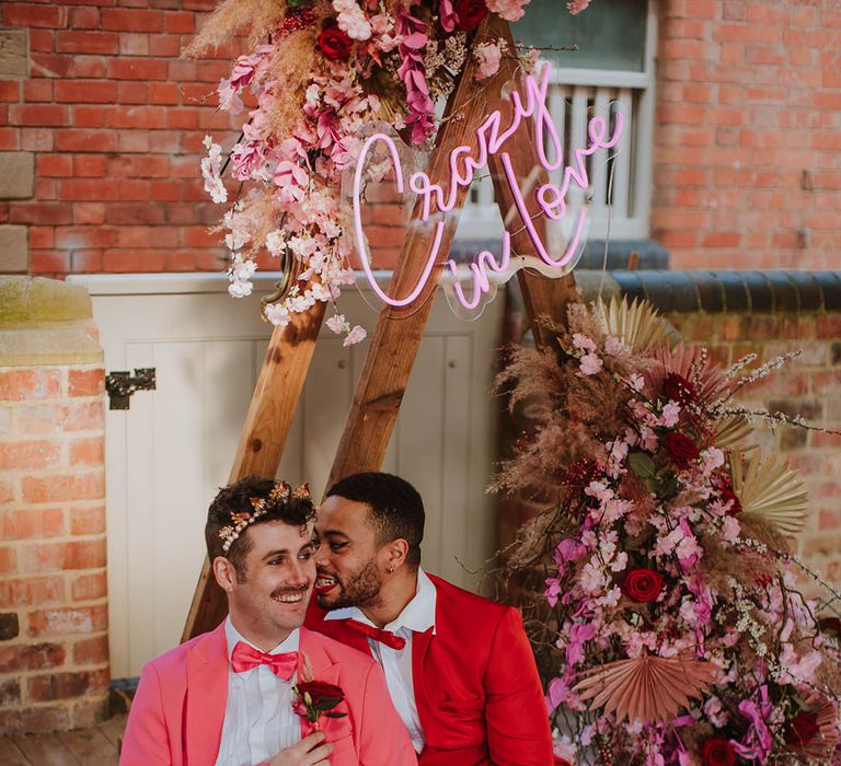 LGBTQI+ couple in pink and red suits sitting in front of a wooden triangle frame altar decorated with a pink neon sign and pink flowers and dried grass floral arrangement 