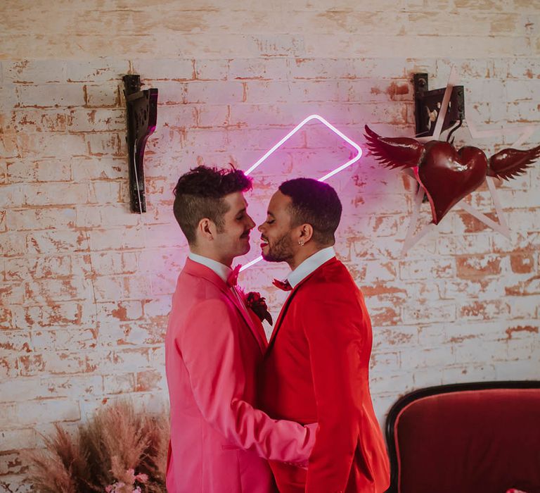 LGBTQI+ couple in pink and red suits standing in front of a pink neon sign at Prestwold Hall Barn wedding venue 