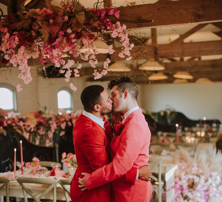 Two grooms in pink and red suits kissing at their Prestwold Hall Barn reception decorated with pink cherry blossom 