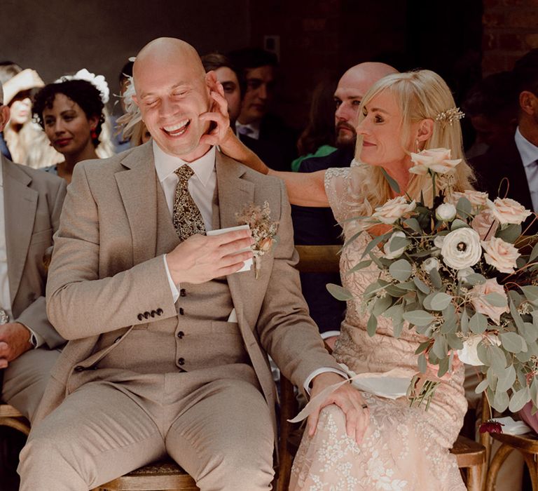 Bride's parents sit together in gold and beige outfits as the mother of the bride squeezes the father of the bride's cheek 